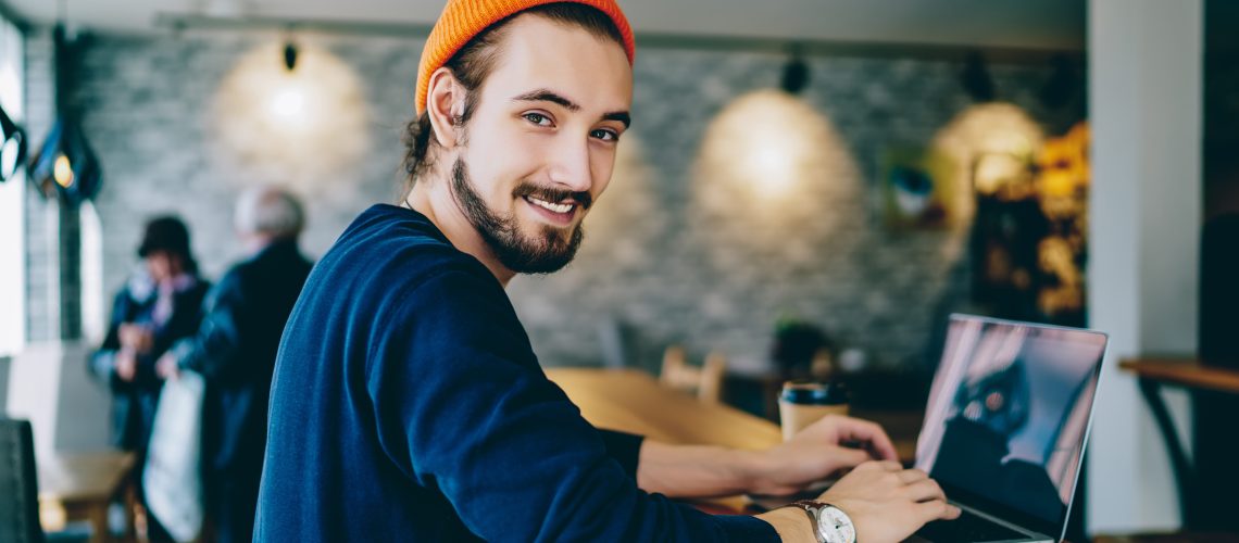 Skilled male it developer looking at camera coding on modern laptop computer working on freelance in cafe interior, portrait of cheerful hipster guy using netbook for e learning satisfied with course